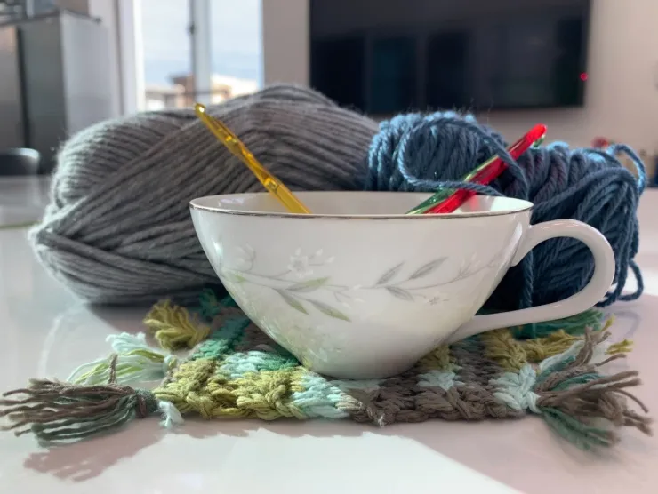 A white tea cup sitting on a green crocheted coaster. In the tea cup are two crochet hooks and two skeins of yarn behind them. 