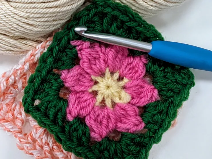 A granny square with a flower-shaped center, shown with a crochet hook and yarn.