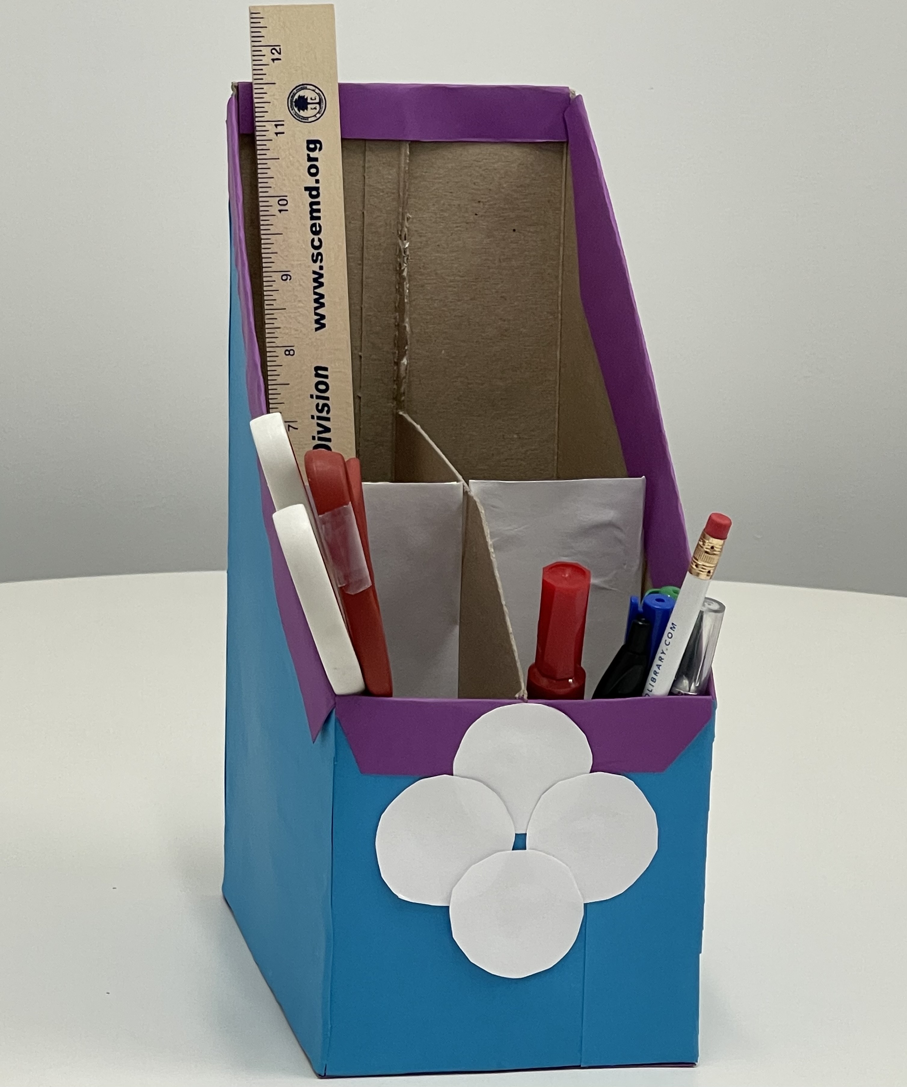 white, purple, and blue desk organizer made out of cardboard; red & red/white handled scissors, red and green markers, white pencil