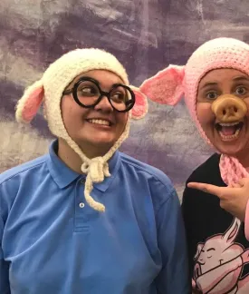Image of two actors.  On the left, the actor wears a long-sleeve blue polo shirt.  They wear round black-framed glasses and a knit cap with sheep ears.  They are looking at the other actor and smiling.  On the right, the actor wears a pink knit cap that looks like pig ears and a snout.  They are also wearing a black t-shirt with a pink long-sleeve shirt underneath.  They are smiling, looking at the camera and pointing at the other actor.  