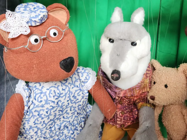 A photo of 4 marionette puppets.  The one on the far left is a Mama Bear.  She is light brown, wears glasses and a blue and white dress with a matching hat.   Next to Mama is a gray and white wolf.  He wears a burgundy patterned shirt and yellow pants.  Beside the wolf is Baby Bear, a very light brown, small fuzzy bear.  On the far right is Papa Bear.  He is dark brown with a lighter belly and muzzle.  He has light blue eyes and wears blue jeans.    