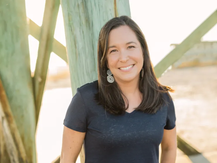 Photo of author Angela May.  She is a white woman in a dark v-neck t-shirt.  She wears light-colored jeans and leans against the structure underneath a pier.  She has shoulder length dark hair and is smiling.  She is also wearing large silver earrings.  