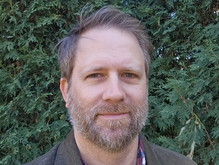 Photo of Matthew Cordell.  He is a white man with short brown hair and a brown and gray beard.  He wears a brown jacket, a plaid shirt and a purple t-shirt.  He is standing in front of greenery and has a slight smile on his face.  