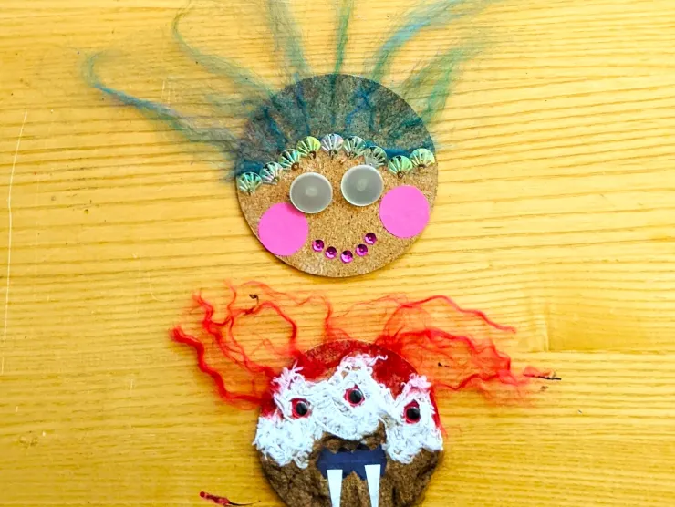 Photo: two cork circles. One is decorated as a mermaid with blue yarn hair, seashell sequins, pearl button eyes, and a pink sequin mouth. The other is a red-yarn-haired monster with white paper fangs, brown yarn fur, and three red googly eyes.